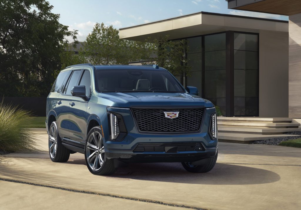 Front ¾ view of the 2025 Cadillac Escalade Sport Platinum in Deep Sea Metallic with a house and greenery in the background.