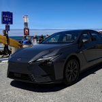 2026 Lexus ES 500e, gray, beach, front 3/4 view, American flag, surfer,